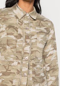 Woman wearing a tan and beige camouflage button-up jacket with frayed edges and front pockets, shown from neck to mid-torso.