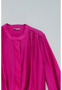 Blusa fucsia a maniche lunghe con colletto rotondo e bottoni frontali nascosti, realizzata in tessuto lucido, posata su uno sfondo grigio.
