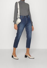 Pull à col roulé gris, jean bleu foncé raccourci et escarpins béiges à bride arrière avec un talon carré. La tenue comprend un sac bandoulière circulaire crème.