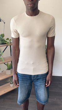 Beige tætsiddende kortærmet skjorte lavet af glat materiale, parret med denimshorts, vist foran en hylde med planter.