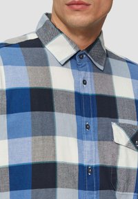 Geruit overhemd met blauwe, grijze en witte tinten. Gemaakt van zachte stof met een knoopsluiting aan de voorkant, een kraag en een enkele borstzak.