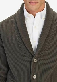 Stickad kofta i mörkbrunt, med sjalkrage, ribbad textur och fyra runda knappar. Bärs över en vit skjorta.