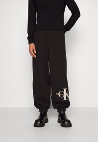 Svarta sweatpants med lös passform, med en framträdande guld Calvin Klein-logotyp på vänster sida. Tillverkade av mjukt material med ribbstickade muddar.
