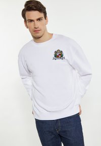 Hvid crewneck sweatshirt lavet af bomuld, med et broderet varsity-logo med et skjold-design og dekorative elementer. Parret med mørke jeans.