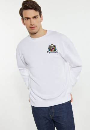 Hvid crewneck sweatshirt lavet af bomuld, med et broderet varsity-logo med et skjold-design og dekorative elementer. Parret med mørke jeans.
