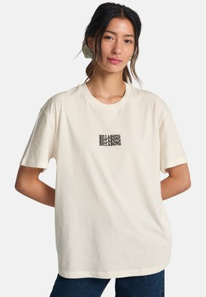 Giovane donna con capelli scuri indossa una T-shirt Billabong color avorio e jeans scuri, in piedi con le mani dietro la schiena contro uno sfondo semplice.