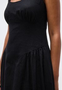 Γυναίκα που φοράει μαύρο αμάνικο φόρεμα με σούρα στη μέση και λεπτομέρεια στη ραφή στο πλάι, εστιάζοντας στη μέση και στο χέρι.