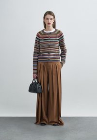 Πλεκτή ζακέτα με ρίγες, στρογγυλή λαιμόκοψη και μπροστινό κούμπωμα με κουμπιά, συνδυασμένη με καφέ παντελόνια φαρδιάς γραμμής, κρατώντας μικρή μαύρη τσάντα.