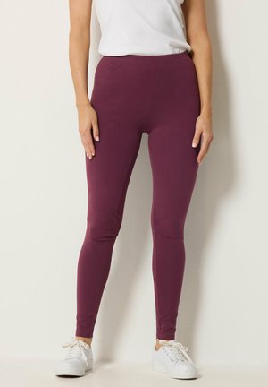 Frau trägt hoch taillierte leggings in Pflaumenfarbe, kombiniert mit weißen Sneakers und einem weißen ärmellosen Oberteil vor einfarbigem Hintergrund.