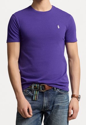 Mężczyzna noszący dopasowany fioletowy t-shirt z krótkim rękawem z małym białym logo na piersi, niebieskie dżinsy, paski w paski i zegarek na rękę.