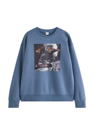 Teenagedreng stående iført blå sweatshirt med tiger skateboard-grafik, sorte jeans og sort-hvide sneakers på ensfarvet baggrund.