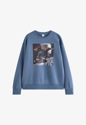 Adolescent debout portant un sweat-shirt bleu avec un motif tigre sur un skateboard, jean noir et baskets noires et blanches sur fond uni.