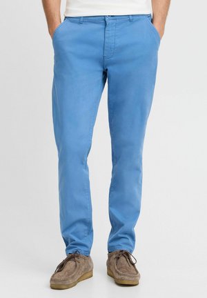 Helleblaue Chinos mit schlankem Schnitt, aus Baumwollmaterial gefertigt, mit seitlichen Taschen und einem vorderen Knopfverschluss. Kombiniert mit braunen Wildlederschuhen.