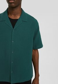 Maglietta a maniche corte color verde acqua realizzata in tessuto testurizzato, con colletto classico e chiusura a bottoni, indossata sopra una maglietta nera.