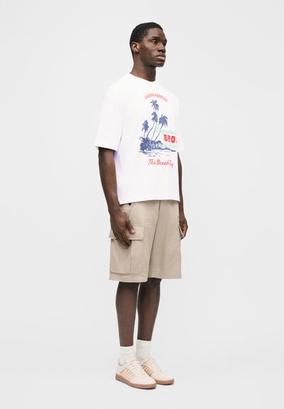 Mann, der ein weißes T-Shirt mit Grafik, beige Cargoshorts, weiße Socken und beige Sneakers vor einem schlichten weißen Hintergrund trägt.