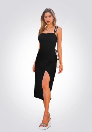 Robe midi noire avec bretelles spaghetti, corsage ajusté, fente sur le côté et nœud ajustable. Portée avec des sandales compensées et un petit sac à main.