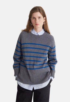 Joven mujer con el pelo largo y castaño que lleva pendientes de aro plateados, un jersey gris con rayas horizontales azules, una camisa blanca y pantalones negros.
