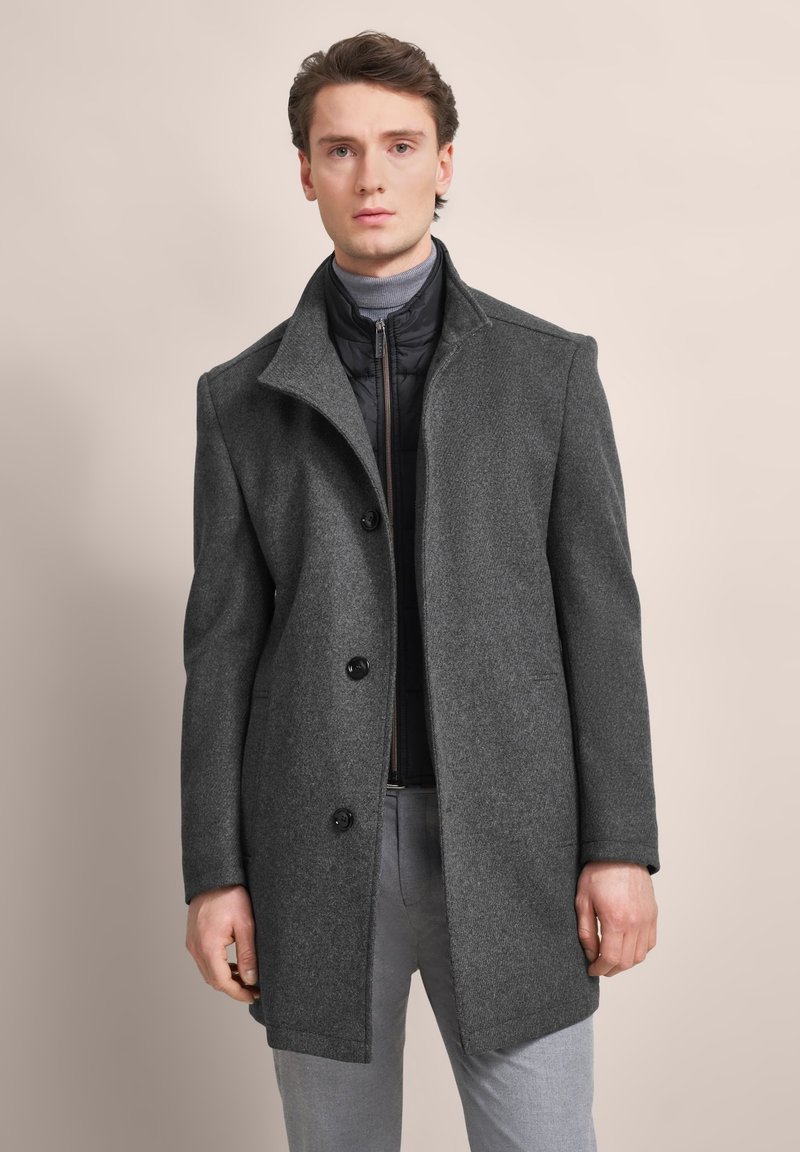 Grauer Wollmantel mit Reverskragen, drei Knöpfen und einer leicht strukturierten Passform. Getragen über einer schwarzen gesteppten Innenjacke.