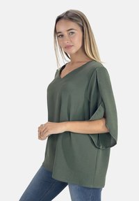 Olijfgroene, loszittende V-hals top met brede mouwen, gemaakt van een gestructureerde stof. Gecombineerd met blauwe jeans.