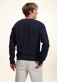 Felpa blu navy realizzata in cotone misto, con maniche lunghe, polsini a costine e scollatura rotonda; indossata con pantaloni chiari.