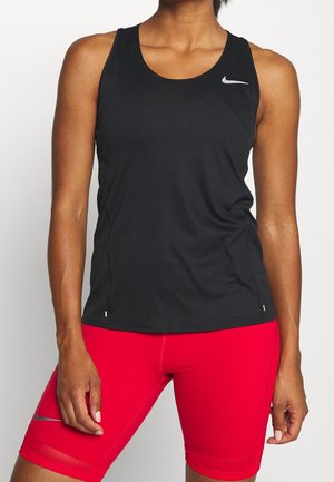 Person iført en sort ærmeløs atletisk top og lyse røde tætsiddende cykelshorts, begge med små reflekterende logoer.