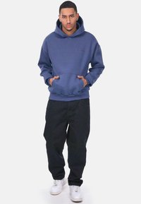 Kapuzensweatshirt in sanftem Blau, mit einer Fronttasche und gerippten Bündchen; kombiniert mit schwarzen lässigen Hosen und weißen Sneakers.