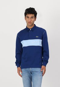 Mornarska polo majica z dolgimi rokavi z svetlo modrim vodoravnim pasom, na katerem je napis "LACOSTE." Ima ovratnik in majhen logotip na prsih.