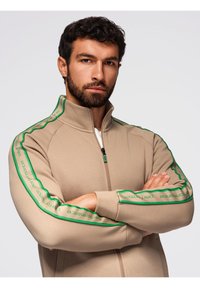 Giacca beige con zip e colletto alto, caratterizzata da strisce laterali verdi con testo e un tessuto testurizzato. Include una tasca con zip.