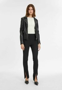 Giubbotto di pelle nera con borchie argentate, top bianco aderente a coste e pantaloni neri con fessure laterali, completati da scarpe nere.