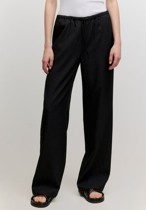 Pantalons larges noirs avec une taille élastique et un cordon de serrage, présentant une texture lisse et une silhouette décontractée. Associés à des sandales plates.