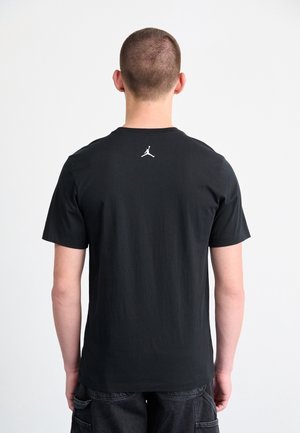 Personne aux cheveux courts portant un t-shirt noir avec un petit logo blanc Jumpman dans le haut du dos, debout devant un fond blanc uni.