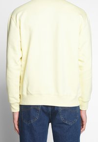Felpa di cotone gialla con polsini e orlo a costine, caratterizzata da un semplice scollo rotondo e una superficie morbida e liscia. Indossata con jeans blu.