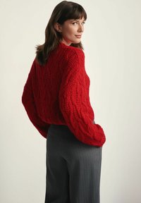 Czerwony sweter na drutach z teksturowanym wzorem ażurowym, o luźnym kroju i opadających ramionach, noszony z ciemnymi spodniami w prążki.