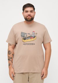 Beige Baumwoll-T-Shirt mit einem Grafikdesign eines Skeletts in einer roten Mütze, das auf einer Holzkiste mit einem Fernseher sitzt und eine Fernbedienung und Pizza hält.