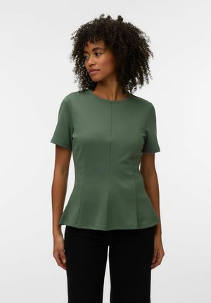 Kvinde med krøllet hår iført en olivengrøn tætsiddende kortærmet top og sorte bukser, der kigger let til siden mod en hvid baggrund.