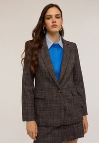 Blazer a quadri marrone con rever a contrasto e due tasche frontali, indossato sopra un maglione blu testurizzato e abbinato a una gonna coordinata.