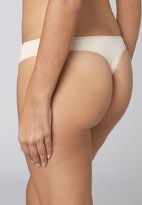 Perizoma in cotone beige con una fascia stretta, texture liscia e delicati bordi smerlati, visto di lato con una mano sul fianco.