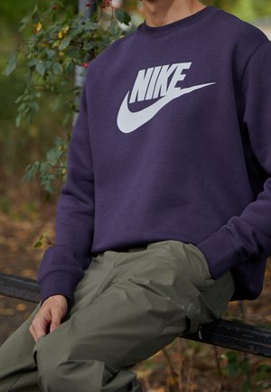 Osoba ubrana w fioletową bluzę Nike z białym logo i oliwkowozielone spodnie, siedząca na ławce na zewnątrz, w tle zieleń.