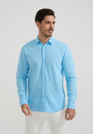 Homme portant une chemise bleu clair à manches longues et un pantalon blanc, debout, les mains détendues le long du corps, regardant vers la droite.