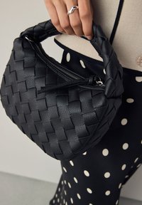 Sac à main en cuir noir tressé de forme arrondie, fermeture éclair et poignée texturée, tenu par une main, à côté d'un vêtement à pois.