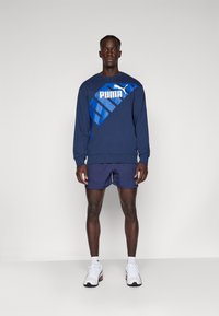 Felpa navy con logo Puma blu audace, abbinata a pantaloni corti navy. Il modello indossa scarpe sportive bianche con accenti neri. Sfondo semplice.