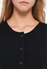 Crna kardigan s okruglim izrezom, s tri crna gumba i teksturiranim pletenim materijalom. Prikaz u blizini ističe uzorak i materijal.