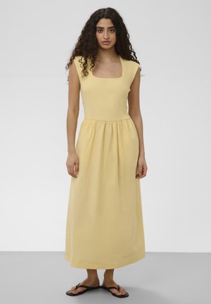 Kobieta o kręconych włosach, ubrana w żółtą sukienkę midi bez rękawów i czarne klapki, stoi na jednolitym szarym tle.