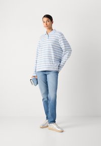 Lichtblauwe polo met lange mouwen met witte en donkerblauwe strepen, gecombineerd met lichtblauwe jeans en zilveren sneakers, terwijl er een blauwe pouch wordt vastgehouden.