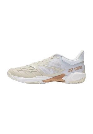 Zapatilla deportiva beige clara con paneles de malla, superposiciones tonales, suela de perfil bajo y logo "YONEX" en bronce en el lateral del talón.