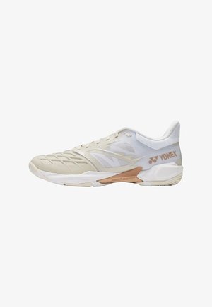 Zapatilla deportiva beige clara con paneles de malla, superposiciones tonales, suela de perfil bajo y logo "YONEX" en bronce en el lateral del talón.
