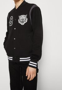 Svart varsityjacka med vit tigerbroderi, randiga muddar och nederkant, samt ett bokstavsemblem. Har grå axeldetaljer och knappar.