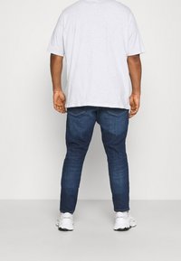 Šedé tričko s krátkým rukávem, kombinované s tmavě modrými úzkými džínami a bílými sportovními botami, zobrazené zezadu na jednobarevném pozadí.