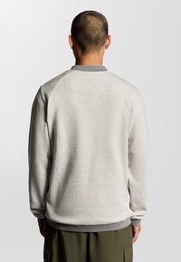Lichtgrijze sweatshirt met ribgebreide grijze kraag en manchetten. Voorwaartse schoudernaden, gestructureerde stof en minimale branding op de halsline.
