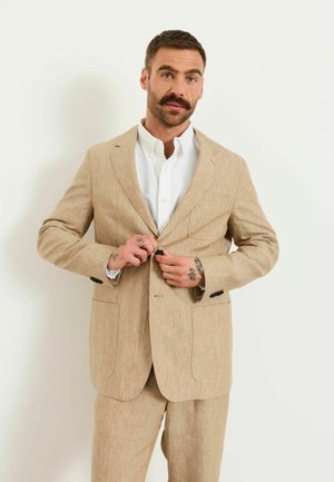Mann mit Schnurrbart und Tätowierungen knöpft beige Leinenjacke über weißem Hemd vor einfarbigem Hintergrund zu